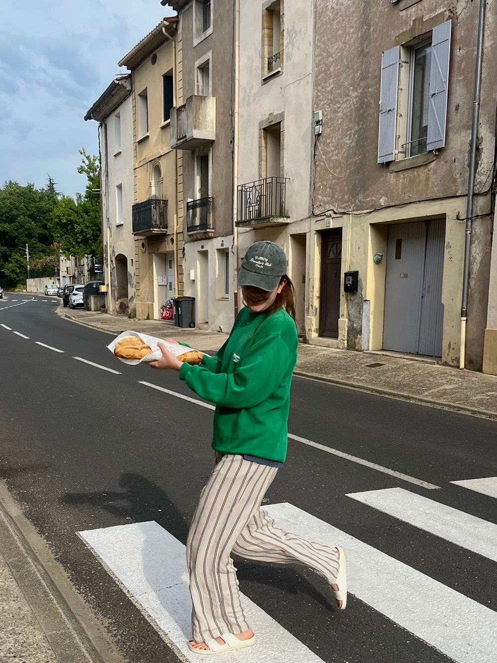 Person in green jacket and cap holding bread on a street with buildings in the background in arrivo italian lifestyle brand