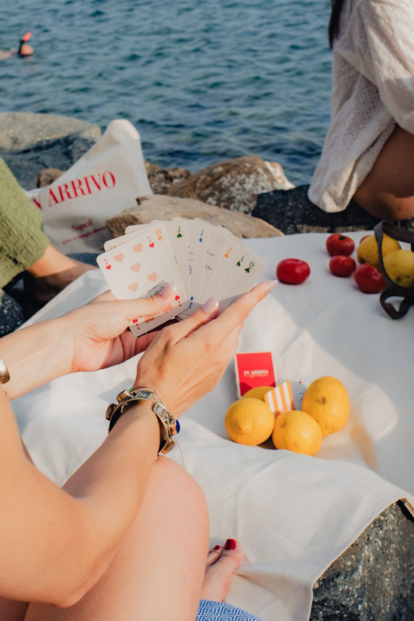 Person playing cards by the sea with fruits and a newspaper.
