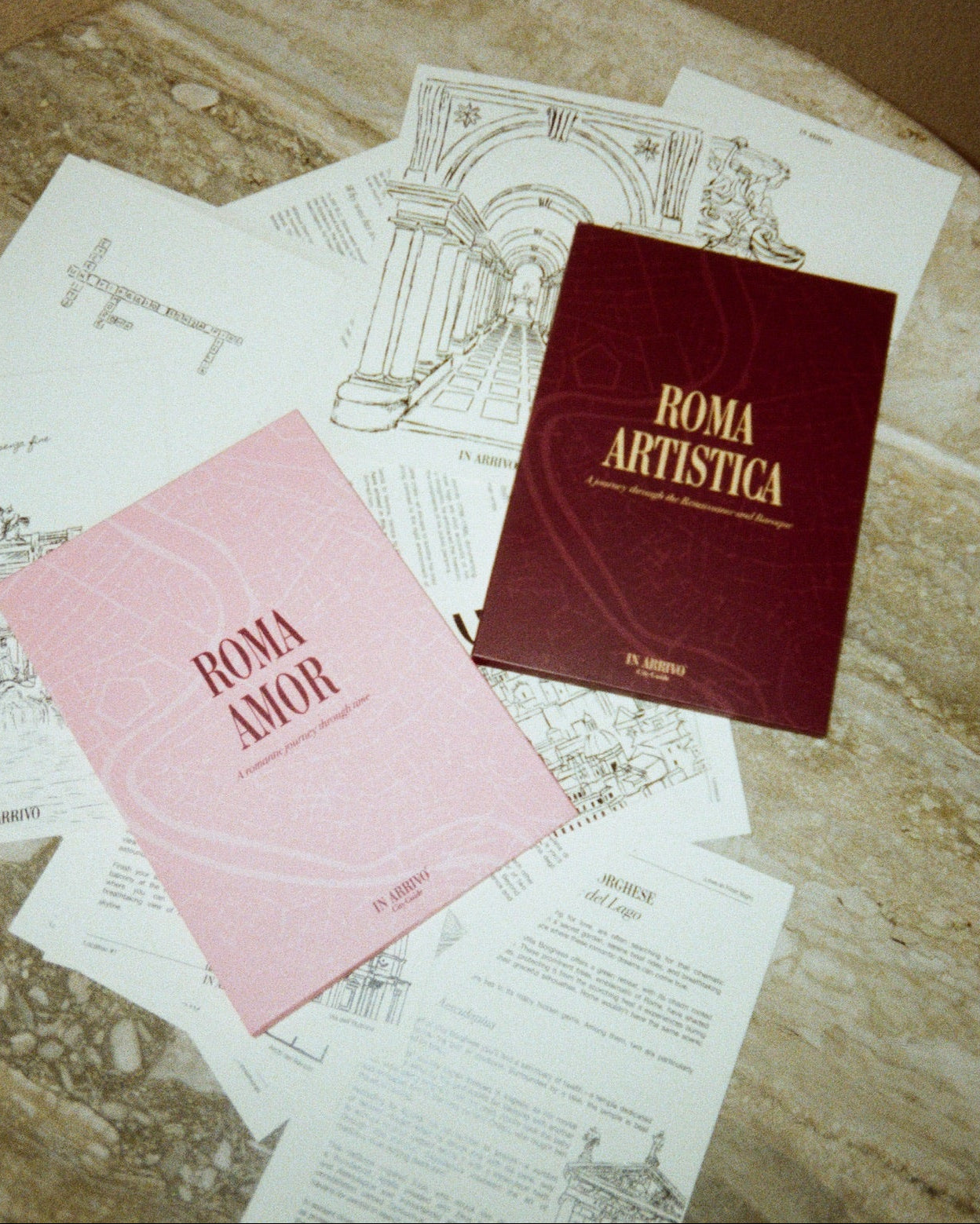 Two books titled 'Roma Amor' and 'Roma Artistica' on a marble surface with architectural drawings.