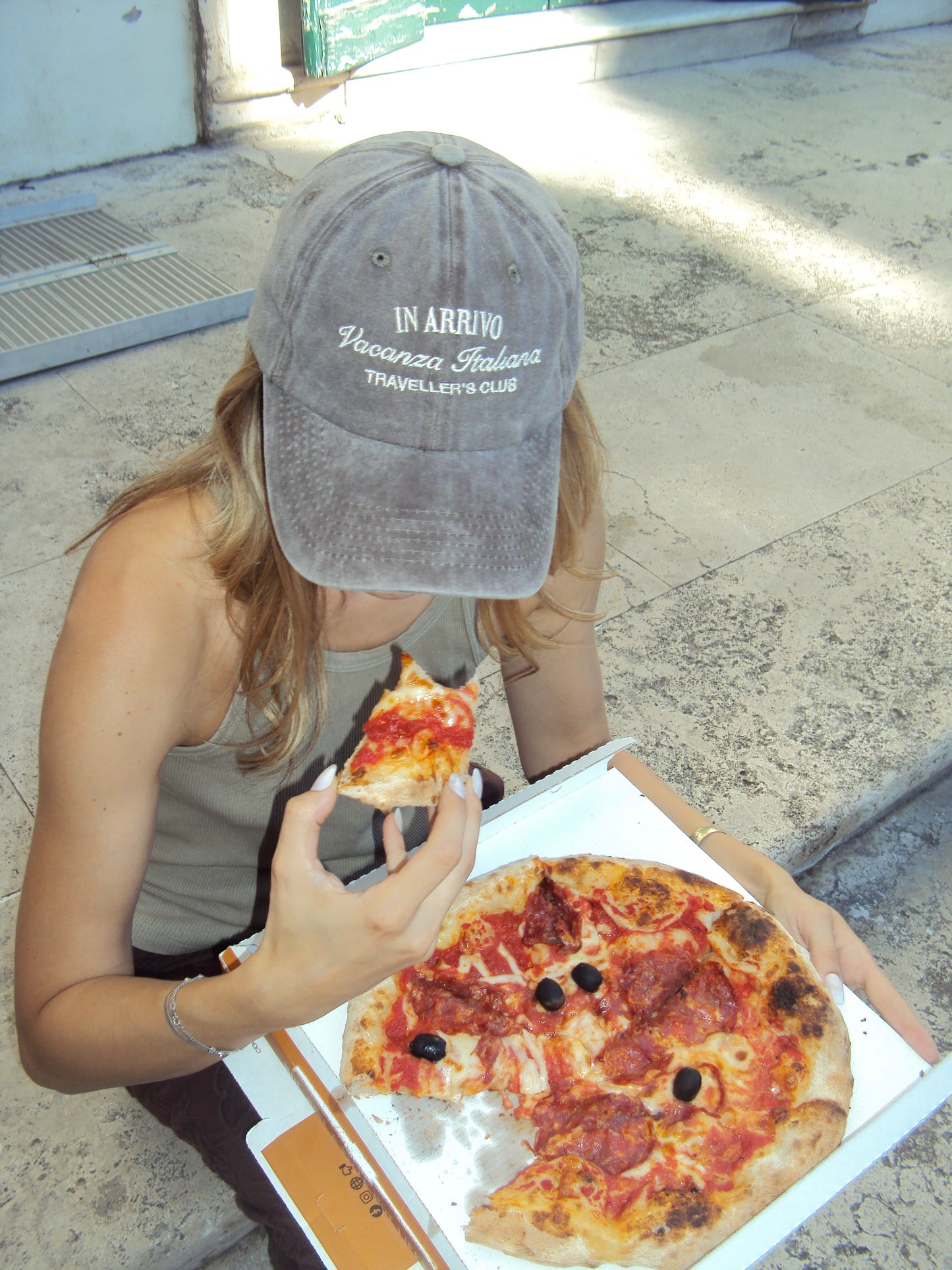 Person eating pizza with a box of pizza on a sidewalk in arrivo italian lifestyle brand brown cap